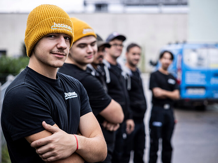 Junger Auszubildender im Vordergrund mit Hackner-Mütze, im Hintergrund weitere Teammitglieder in Reihe