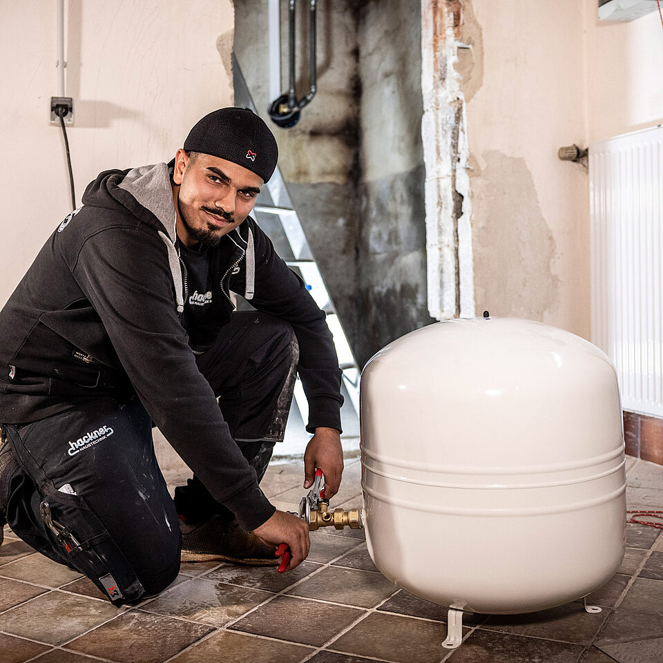 Techniker kniet auf dem Boden und schließt ein weißes Ausdehnungsgefäß an eine Heizungsanlage an