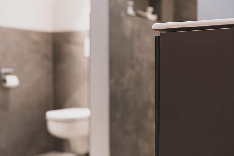 Teilansicht eines Badezimmers mit Fokus auf dunklem Waschtischunterschrank, WC im Hintergrund unscharf