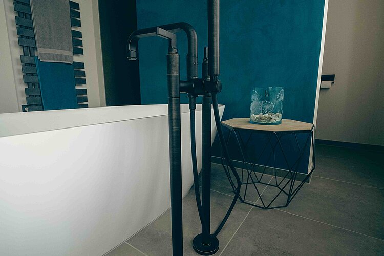 Detailaufnahme eines Badezimmers, die eine moderne, freistehende Badewannenarmatur in Schwarz zeigt. Der Armaturenkörper steht auf dem grauen Boden. Im Hintergrund ist eine leuchtend türkisfarbene Wand zu sehen und daneben ein Handtuchwärmer mit einem blauen Handtuch.