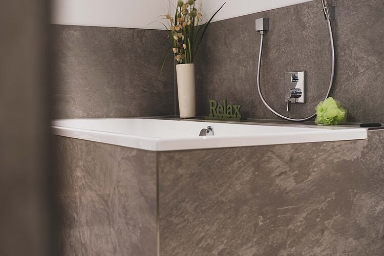 Weiße Badewanne mit dunkler Natursteinverkleidung, Wandarmatur und Dekovase im Hintergrund