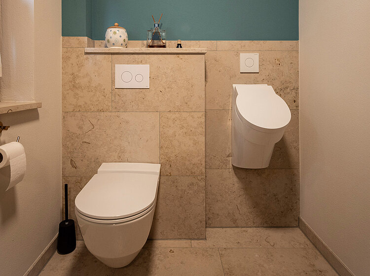Kleines Badezimmer mit wandhängendem WC und Urinal vor beigen Fliesen und türkisfarbener Wand