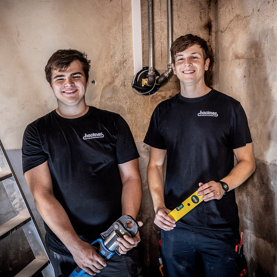 Zwei Auszubildende stehen vor Wand mit Rohrinstallation, halten Wasserwaage und Presszange