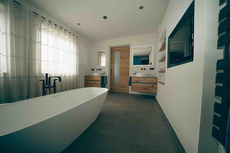 Alt-Text Ein modernes Badezimmer mit einer freistehenden, weißen Badewanne im Vordergrund. Im Hintergrund befinden sich ein Doppelwaschtisch mit Holzschränken und runden Waschbecken.