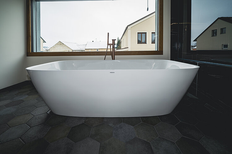 Freistehende weiße Badewanne mit Ausblick durch großes Fenster, elegante Armatur in Bronzeoptik