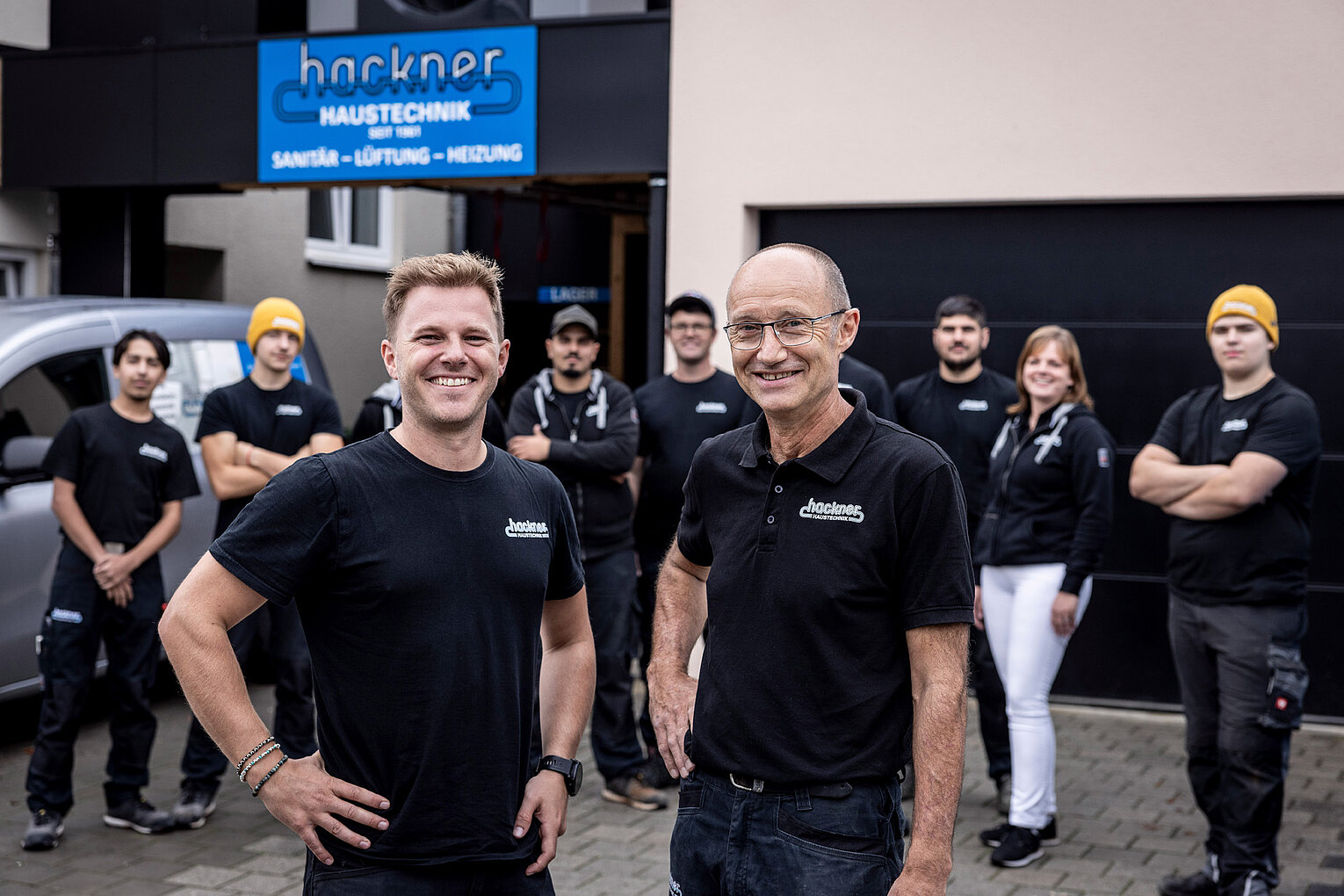 Zwei Führungskräfte von Hackner Haustechnik lächeln in die Kamera, das Team steht im Hintergrund